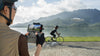 Person in activewear gear operating a drone with remote controller to film another cyclist on a bike with lake and mountainous background