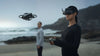 Woman wearing FPV goggles and operating the DJI Avata 2 grey drone with a man standing in the background on a beach