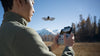 Man using a remote controller to operate a DJI Neo grey drone with a natural mountainous setting in the background