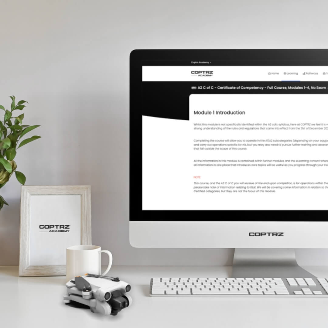 Computer monitor displaying the online training portal, on a desk with a drone, mug, plant and photo frame.