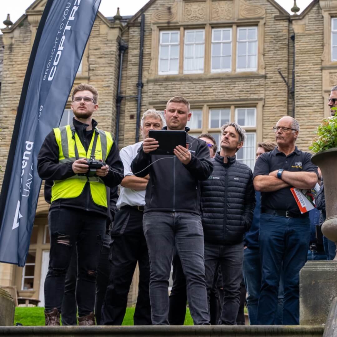 Group of people standing outdoors operating a drone and watching the screen on a controller with a building in the background