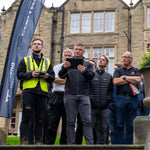 Group of people standing outdoors operating a drone and watching the screen on a controller with a building in the background