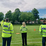 Three police officers in high-visibility jackets operating drone on a grassy field with trees and houses in the background