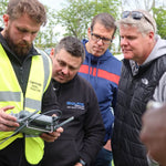 Four men outdoors, three of whom are looking at a remote controller held by one of them that is a trainer