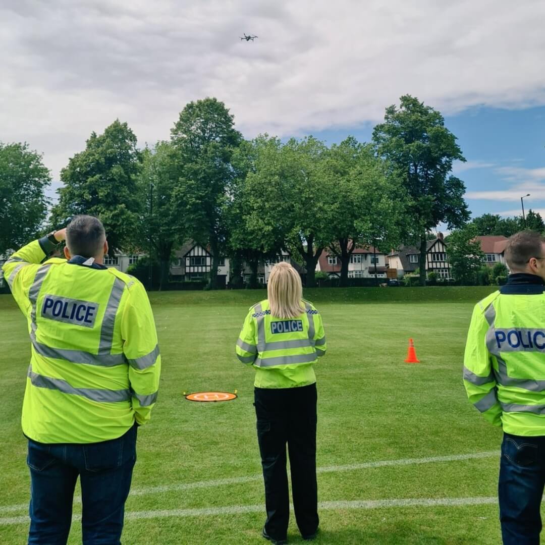 Three police officers in high-visibility jackets operating a drone on a grassy field with trees and houses in the background