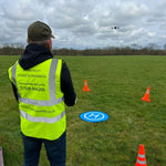 Person in a high-visibility vest operating a drone in an open field with cones and a blue and white landing pad on the ground