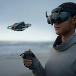 Person using an FPV headset and controller on a beach with an Avata 2 drone flying in the background