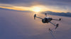 Unmanned aerial vehicle flying over a snowy landscape with mountains at sunset.