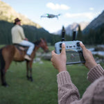 A person using a remote control to operate a drone with a scenic background of mountains and a horse