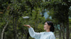 Woman in a light blue jacket standing in a forest with DJI Neo drone hovering over her hand