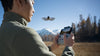 Man using a remote controller to operate a DJI Neo grey drone with a natural mountainous setting in the background
