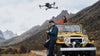 A man and woman operating a grey drone near their yellow car with a mountainous background