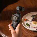Hand holding Osmo Pocket 3 black handheld camera with a blurred plate of food in the background
