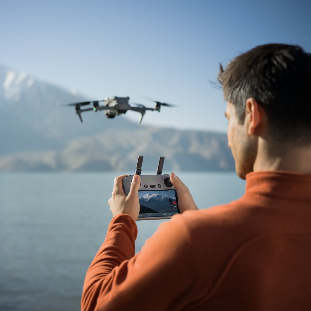 Person operating a DJI Air 3S drone with an RC 2 controller over a scenic lake and mountain backdrop