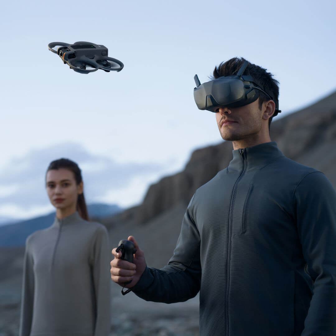Person wearing DJI FPV goggles and holding a remote control with another person in the background