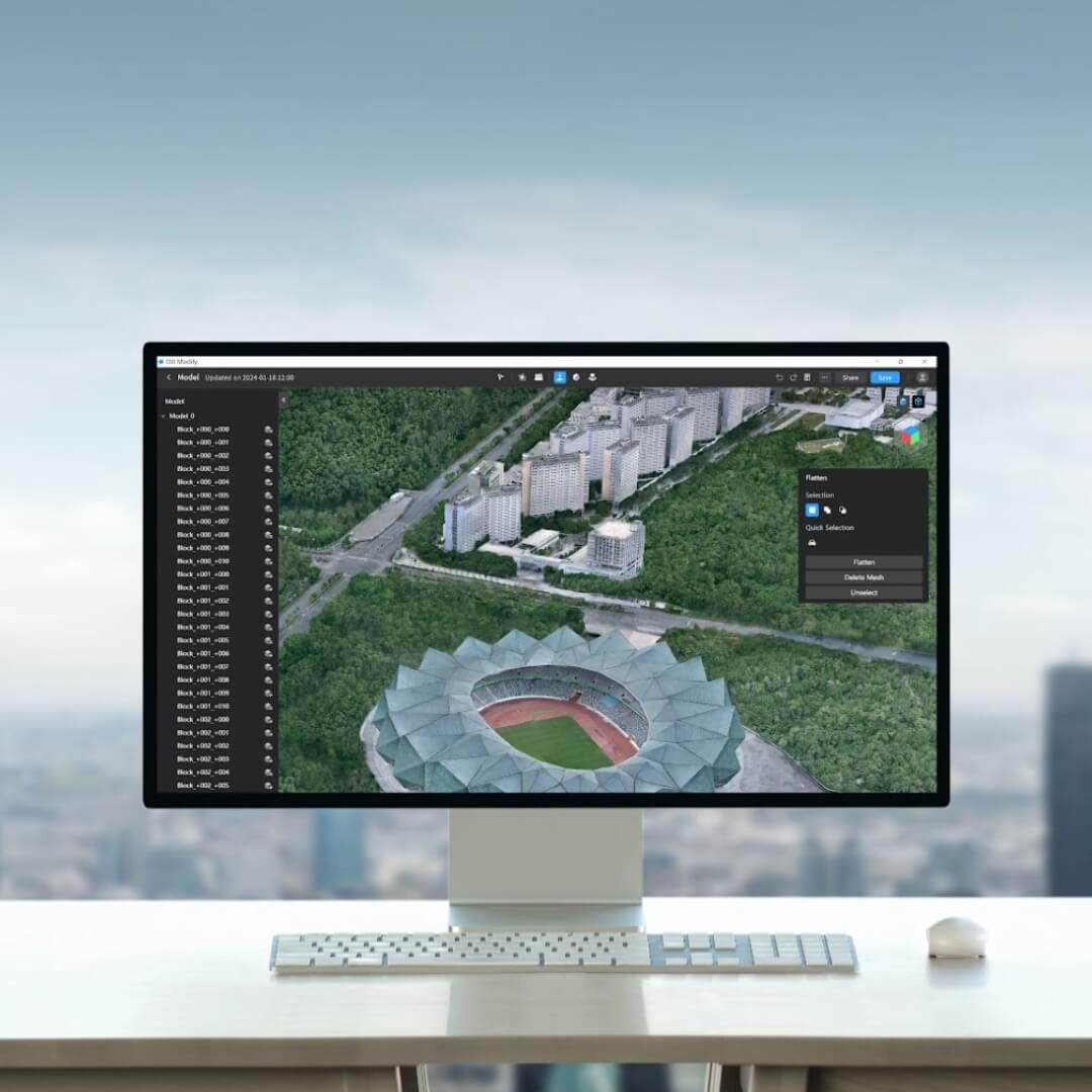 Computer monitor displaying a 3D architectural model of a stadium and surrounding area on a desk with a blurred cityscape background