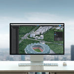 Computer monitor displaying a 3D architectural model of a stadium and surrounding area on a desk with a blurred cityscape background