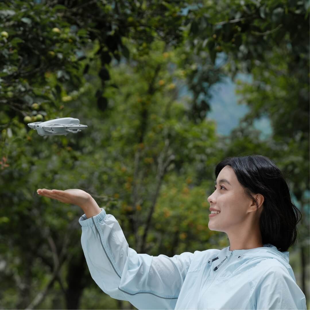 A woman holding her hand below a flying drone in front of a green, leafy background