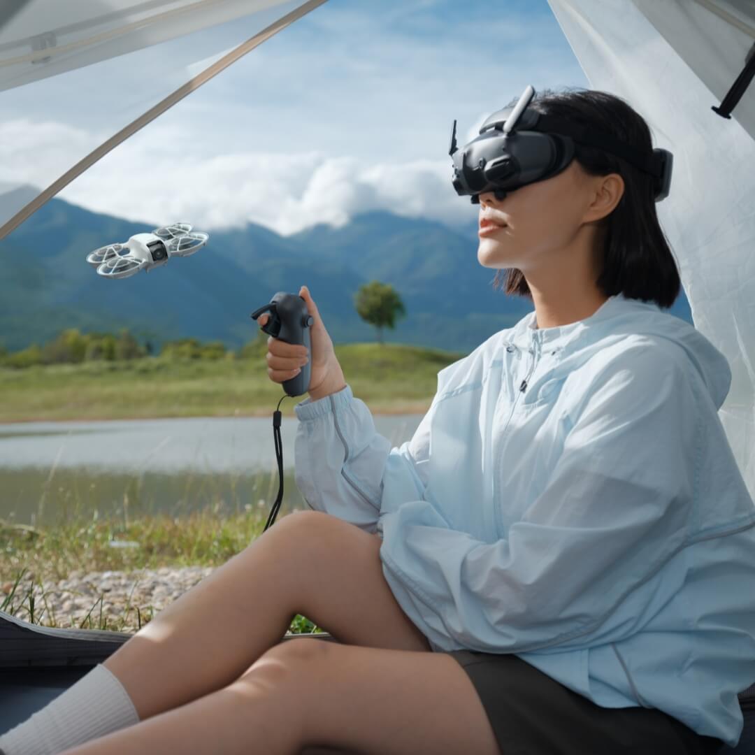 A woman sat in a tent wearing FPV goggles and holding a controller, with a drone flying near them set against a scenic outdoor background